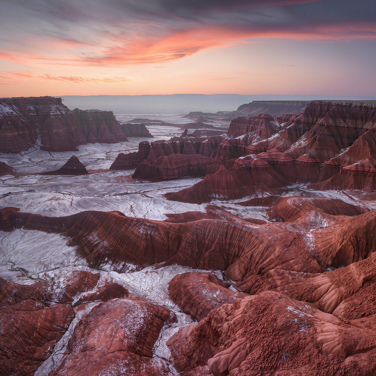 A red-sandstone badlands plateau beneath clearing winter clouds at sunrise, overnight frost silvering the gullies; cracked clay and wind-smoothed rock lead the eye across layered mesas fading into pale distance. — Kling v3