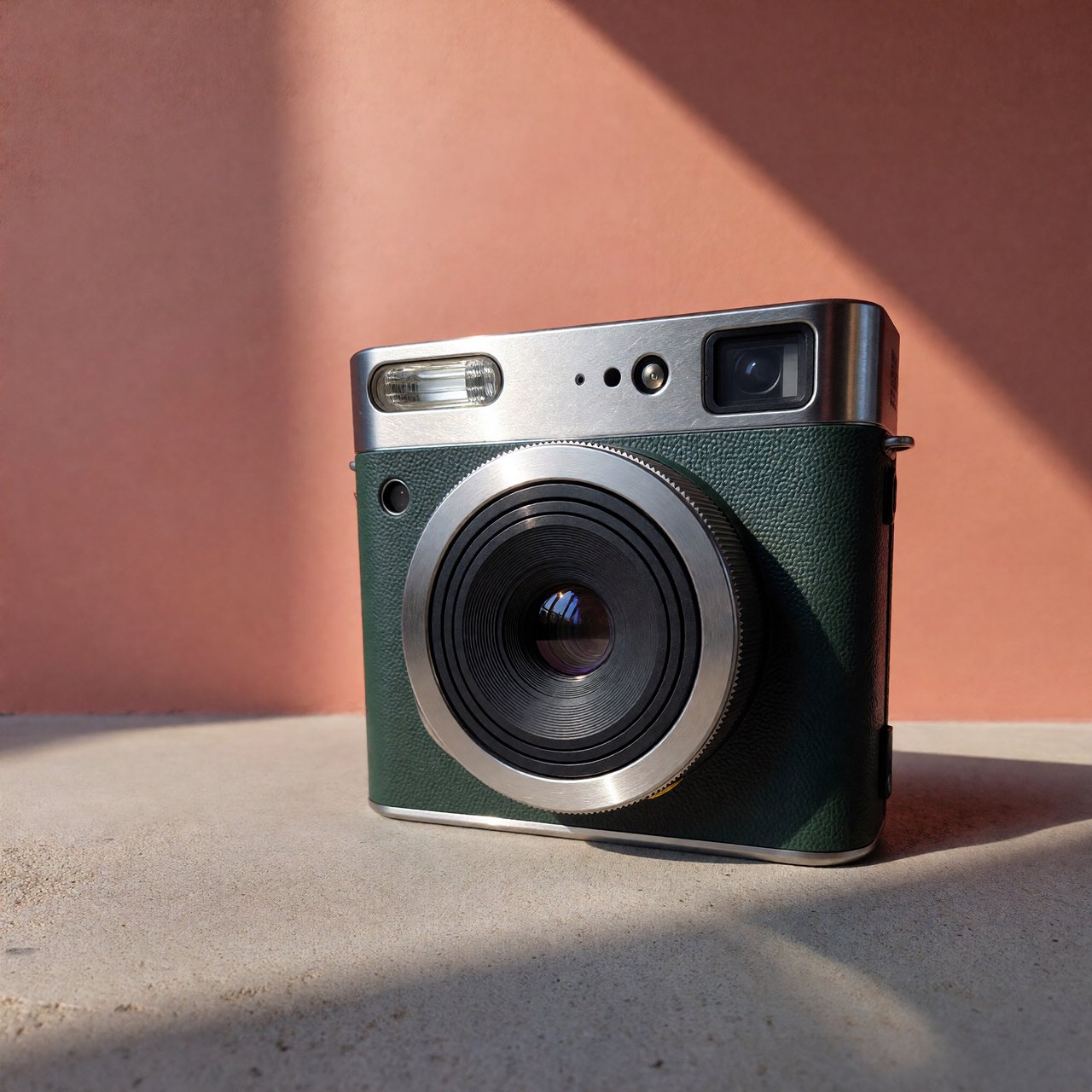 Commercial product photograph of an unbranded compact instant camera in matte forest-green polymer with brushed steel details, resting on a pale concrete surface against a warm peach background, illuminated by broad diffused studio light and a soft rim light. — ImagineArt 1.5 Pro
