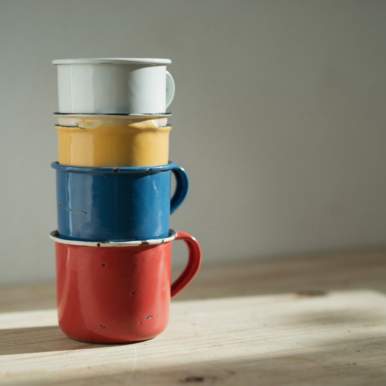 Exactly four enamel camping cups form a vertical tower on a pale wooden shelf, bottom to top: red, yellow, blue, white; all handles face right. Soft morning light, nothing else. — Ideogram v4