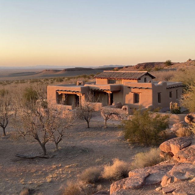 Beyond dry orchard rows, a Pueblo Revival adobe house settles into the desert foothills, with timber lintels and a muted clay roof; warm late-afternoon light, viewed from a rocky rise, reveals clean geometry and credible construction. — ImagineArt 2