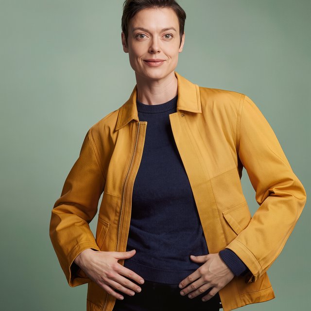 Fashion portrait of an adult nonbinary person in their late twenties wearing a structured saffron-yellow canvas jacket over a fine-gauge navy merino top, hands resting on the jacket hem. Their bright, self-assured expression contrasts with a softly lit sage-green backdrop. — Kling v3