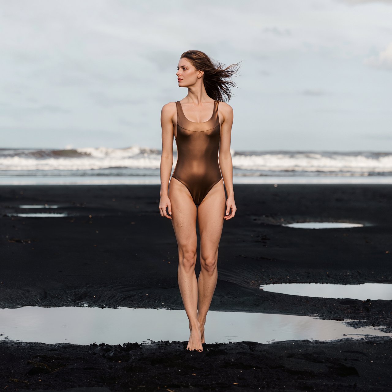 Editorial glamour photograph of an adult woman in a bronze one-piece swimsuit, standing at ease on a quiet black-sand beach beside tide pools, flattering overcast light softly shaping the scene, wind-lifted hair, natural gaze toward distant surf. — Kling v3