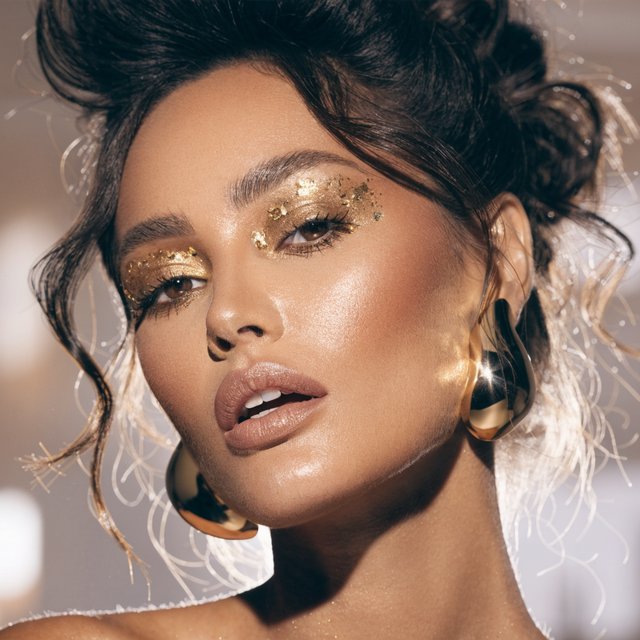 Beauty close-up of an adult woman with bronzed skin and gold eye makeup, bare shoulders, studio backlight halo, high-end campaign look. — Recraft v4
