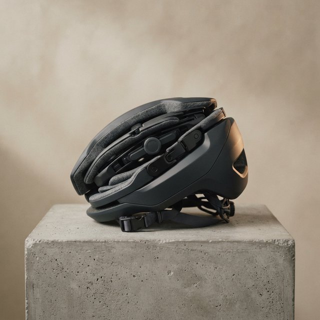 Commercial product photograph of an unbranded folding bicycle helmet made from matte charcoal polymer with soft gray padding, resting on a honed concrete plinth against a muted sand background, illuminated by a broad overhead softbox and gentle rim light. — Nano Banana 2 Lite