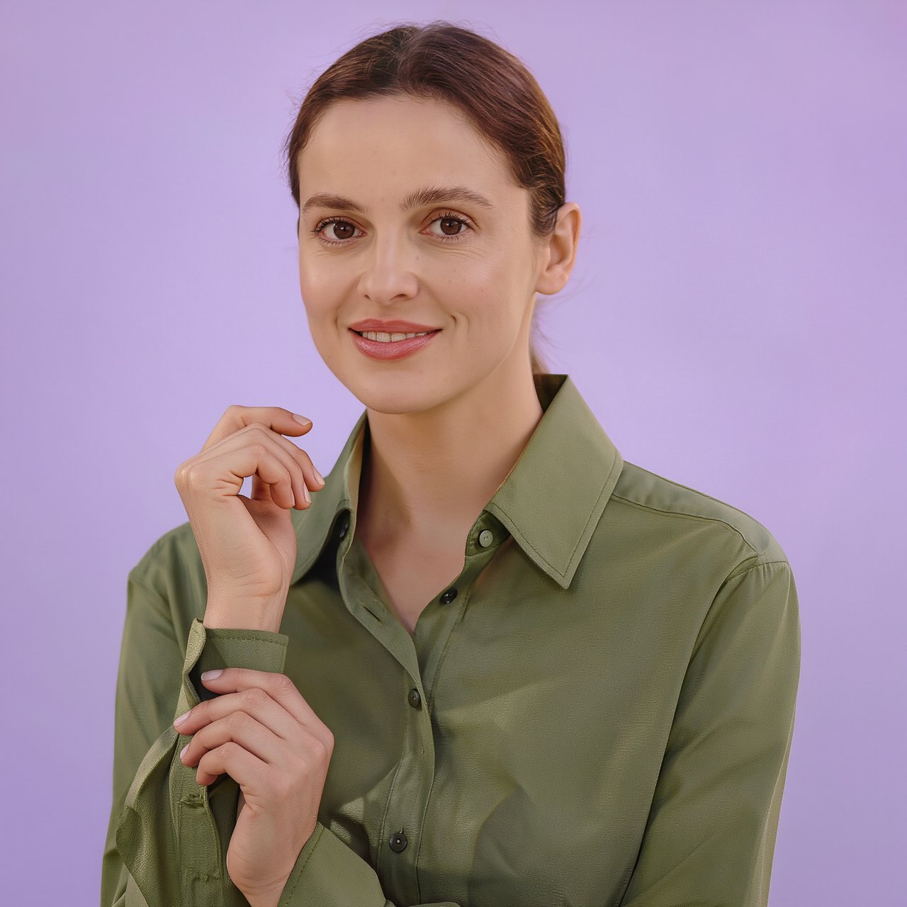 Portrait photograph of an adult woman in her late 30s wearing a moss-green cotton poplin shirt, fingers lightly turning its rolled cuff. Her calm, observant expression faces the camera against a chalky lavender backdrop, lit by soft diffused skylight. — Kling v3