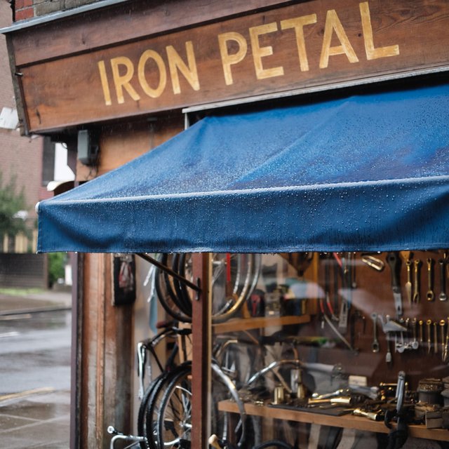 Rain beads on a bicycle repair shop’s blue awning, with wheels and brass tools visible behind the glass; “IRON PETAL” is lettered in warm yellow enamel capitals across the wooden fascia. — ImagineArt 2