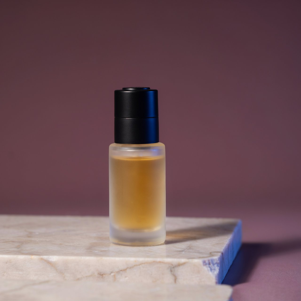 Under a broad diffused key and cool edge light, an unbranded frosted-glass serum bottle displays clear golden oil against a muted plum background, resting on honed pale travertine with its blank surface free of lettering. — Ideogram v4