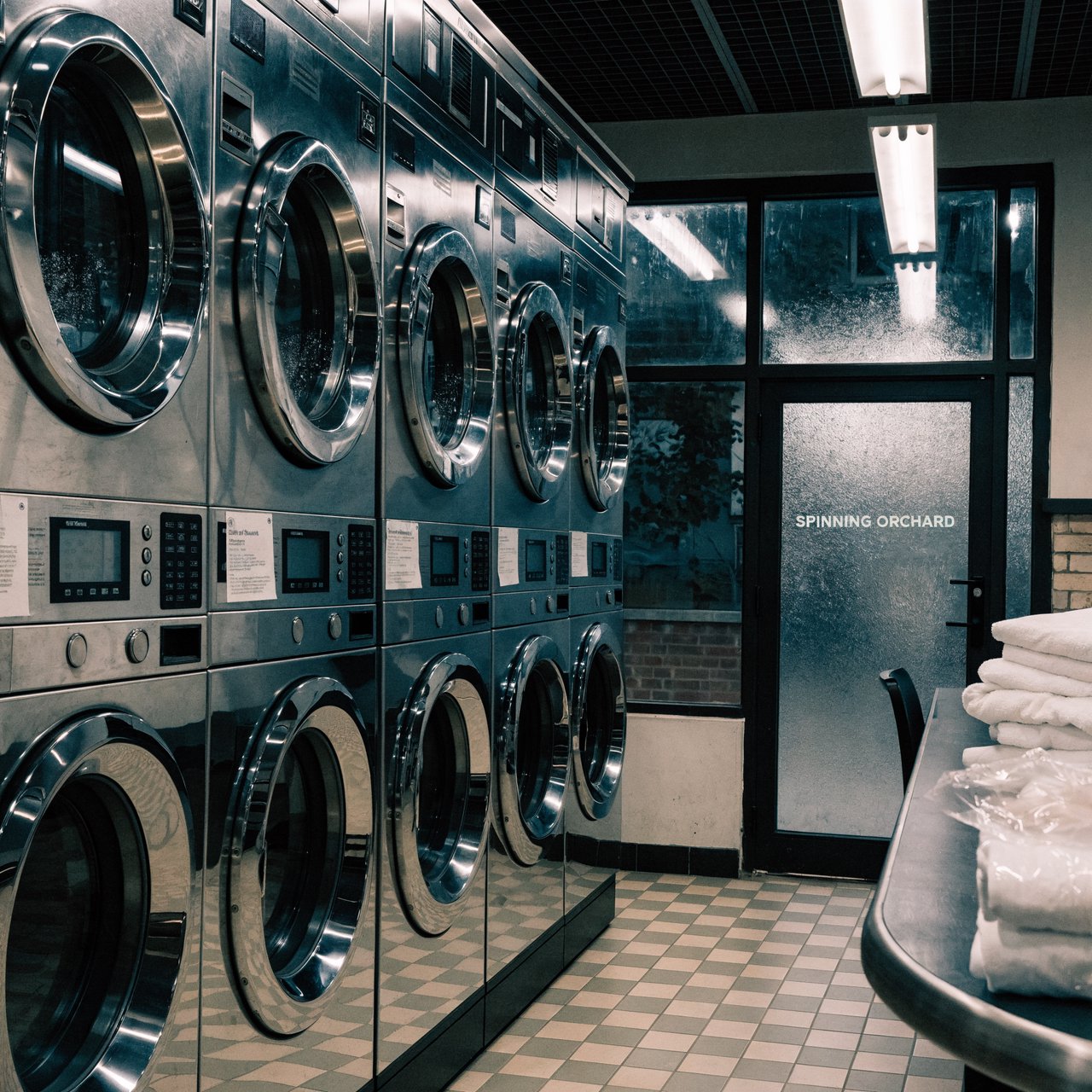 Inside a quiet neighborhood laundromat, chrome machines and folded linen glow beneath cool ceiling lights; "SPINNING ORCHARD" is printed in pale mint letters on the frosted glass entry door. — Recraft v4 Pro
