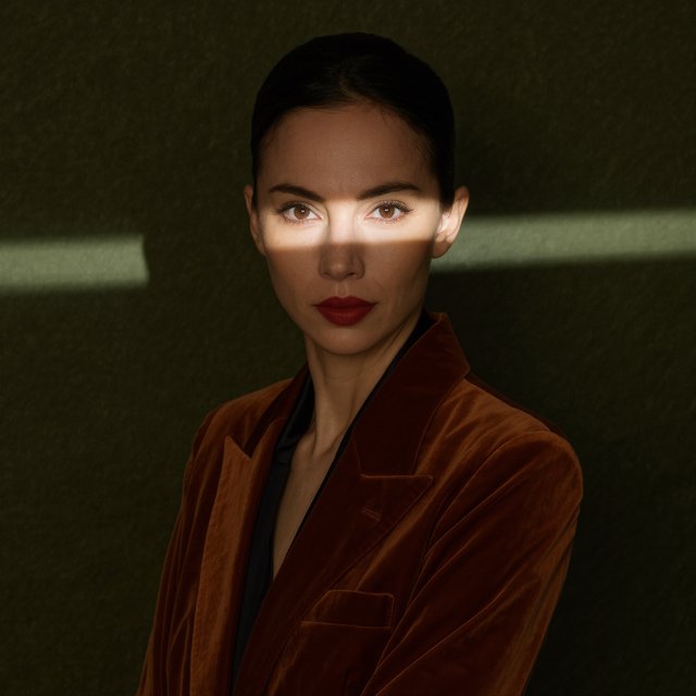 Her guarded gaze catches a solitary beam in a low-key fashion portrait: an adult woman aged 27–35 wears a rust-colored velvet blazer, the rest of her face falling into shadow against a deep olive backdrop. — Kling v3 Omni