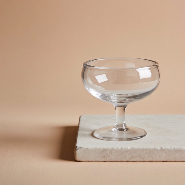 Beside a pale limestone block, an unbranded handblown cocktail coupe in clear soda-lime glass catches crisp window-shaped highlights; its curved bowl bends a muted apricot backdrop under broad diffused key light and narrow cool rim lighting, commercial product image, no lettering. — Krea 2 Medium