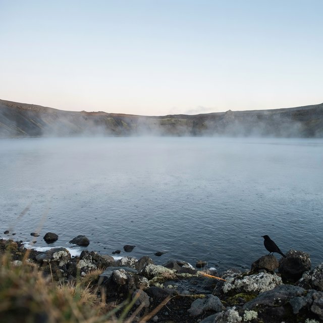 At first light, a rain-cleared volcanic caldera lake lies beneath thinning fog; pumice grains and damp lichen sharpen the near shore, while dark cinder slopes step back toward a distant rim. — Ideogram v4