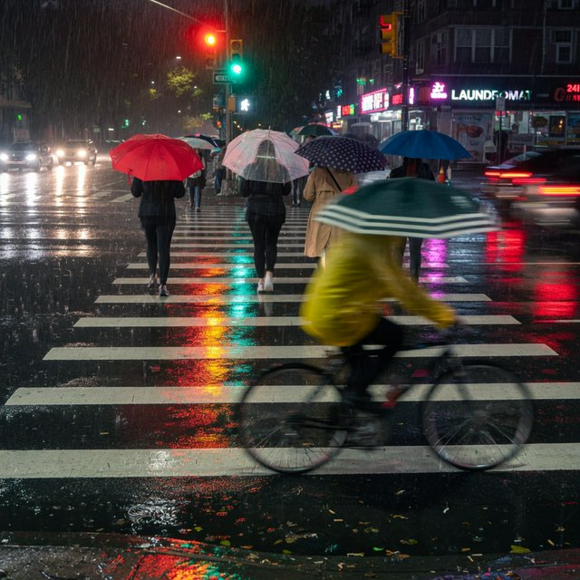 A rain-soaked city crosswalk at night seen from across the street, umbrellas, reflections of traffic lights on wet asphalt, slight motion blur on a passing cyclist. — Grok Imagine Image Quality