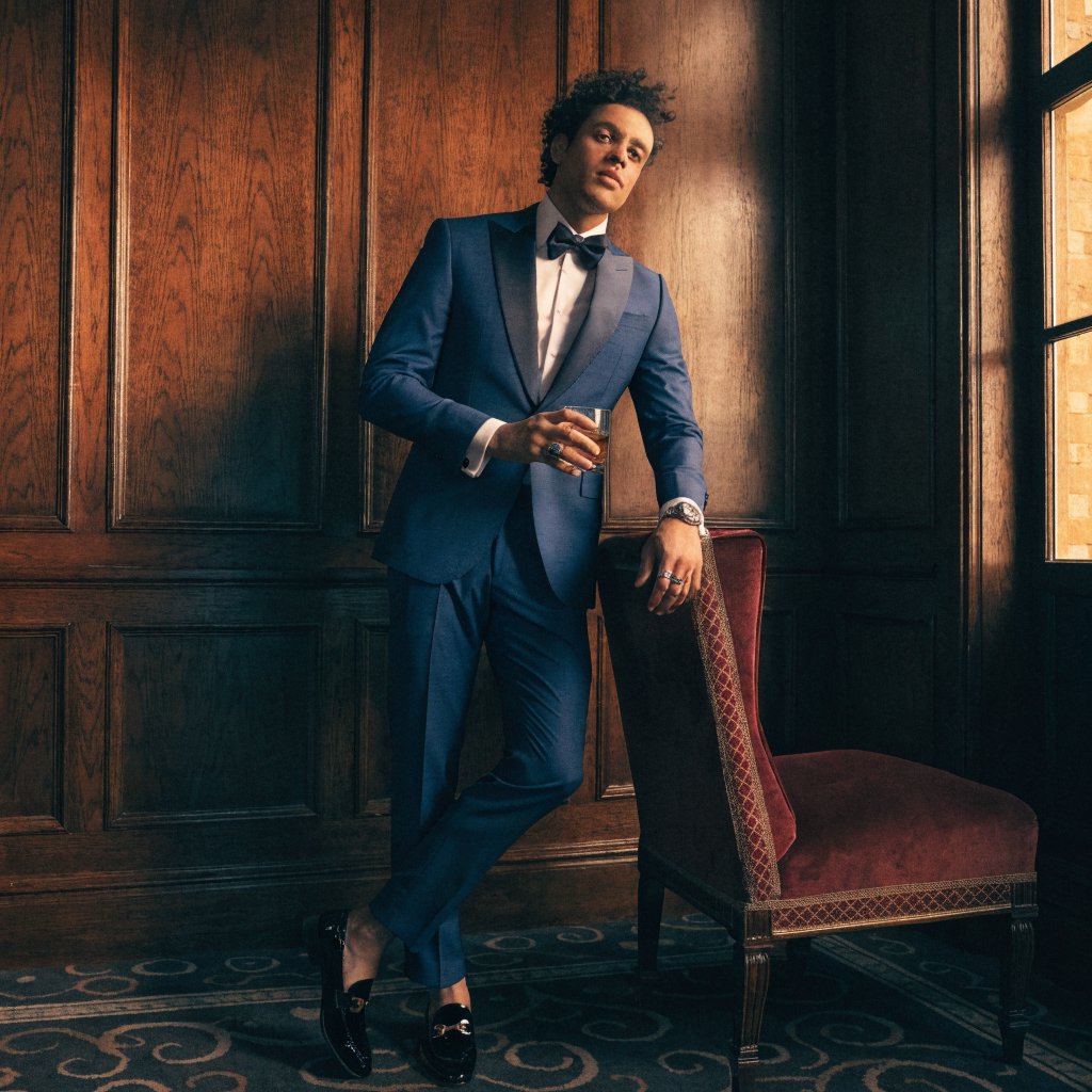 Editorial glamour photograph of an adult man in a midnight-blue tuxedo, standing naturally beside a velvet chair in a quiet hotel lounge, flattering amber window light, relaxed expression, polished magazine composition. — Recraft v4