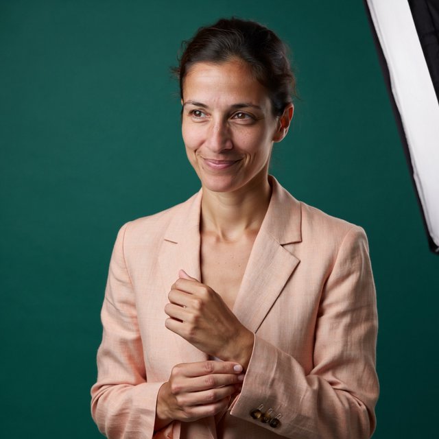 Portrait photograph of an adult woman in her early 30s wearing a pale apricot linen blazer, her hands gently smoothing its cuff. She looks quietly delighted against a deep teal paper backdrop, illuminated by soft, angled studio light. — ImagineArt 2