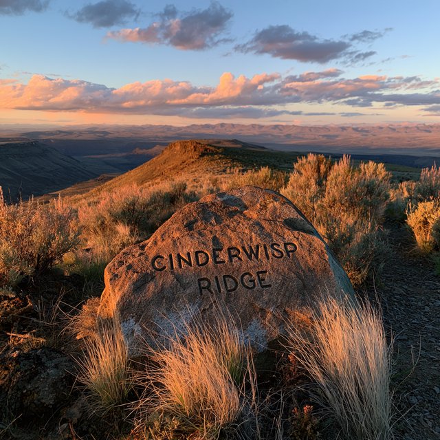 At a high desert canyon overlook, a weathered granite trail marker rises among sage and pale grasses; "CINDERWISP RIDGE" is deeply carved across its broad face, the cut letters catching slanting amber light. — Midjourney V8.2