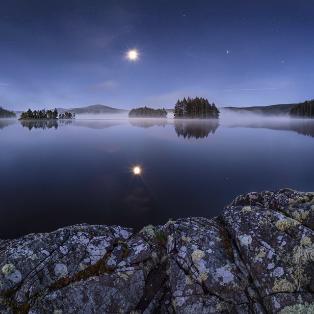 An expansive boreal lake beneath a clear, cold midnight sky, pale moonlight catching thin mist above dark water; spruce-covered islands recede toward distant ridges, while lichen and cracked granite sharpen the near shoreline. — Kling v3
