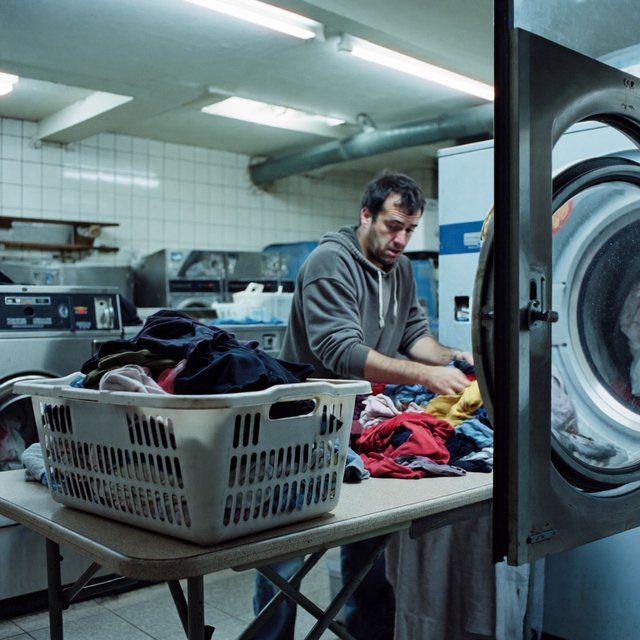 Candid documentary photograph of an adult sorting laundry in a neighborhood laundromat, cool fluorescent light, 35mm lens with moderate depth of field, lint specks floating near a tumbling dryer door. — ImagineArt 2