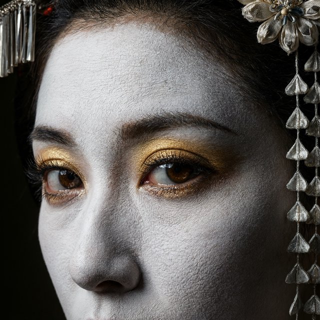 Extreme close-up macro photography of a Geisha's face. The focus is on her eyes, revealing intense emotion and hyper-realistic detail. Her skin is covered in traditional white 'Oshiroi' makeup, showing subtle, realistic cracking and skin texture. Dramatic Chiaroscuro lighting with deep shadows and a single soft light source highlighting the shimmer in her golden eyeshadow and the wetness of her eyes. Ornate kanzashi (hair ornaments) visible in the blurred periphery with intricate silk flowers and dangling silver petals. Cinematic composition, shot on 100mm macro lens, incredibly detailed eyelashes and iris patterns. — Model 18