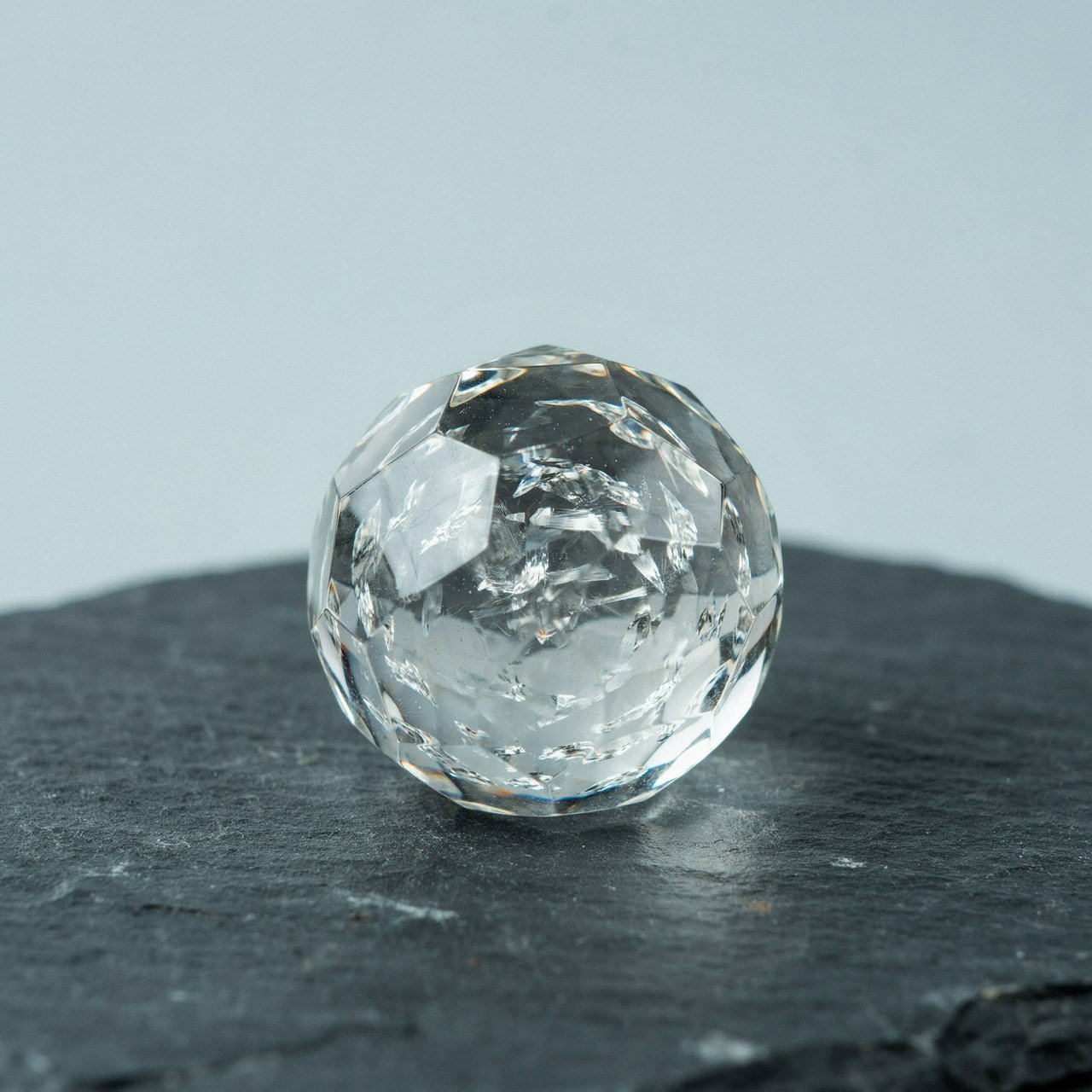 An unbranded, unmarked faceted optical-glass paperweight rests on charcoal slate against a pale blue-gray background; broad diffused studio light and a narrow side highlight reveal crisp internal refractions and bright edge reflections. — Ideogram v4.5