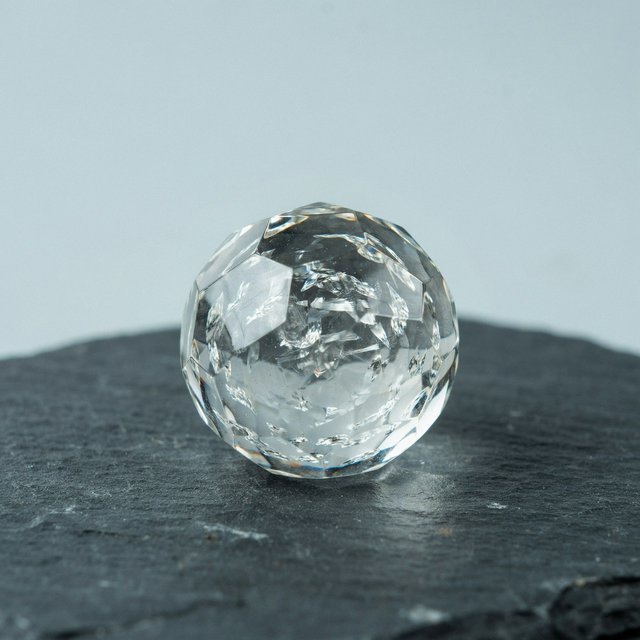 An unbranded, unmarked faceted optical-glass paperweight rests on charcoal slate against a pale blue-gray background; broad diffused studio light and a narrow side highlight reveal crisp internal refractions and bright edge reflections. — Ideogram v4.5