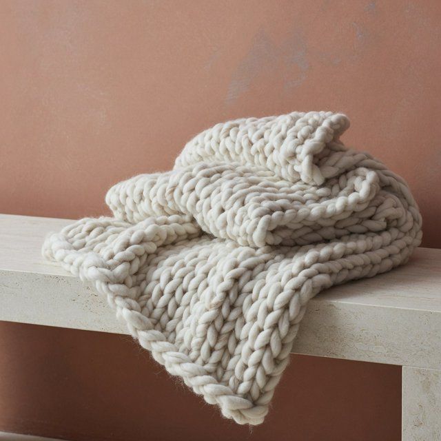 Soft folds of an unbranded chunky-knit wool throw rest on a pale travertine bench; its looped surface catches broad diffused key light and gentle side fill against a muted terracotta studio background, forming a clean commercial product image with no lettering. — Model 6