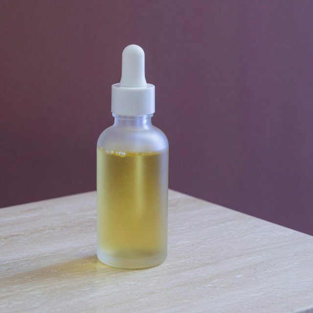 Under a broad diffused key and cool edge light, an unbranded frosted-glass serum bottle displays clear golden oil against a muted plum background, resting on honed pale travertine with its blank surface free of lettering. — Grok Imagine Image Quality