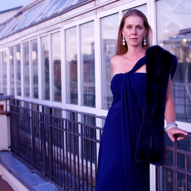 Editorial glamour photograph of one adult woman in a sapphire one-shoulder evening gown, leaning naturally against a rooftop conservatory railing at dusk, flattering lavender light, glass reflections, composed expression, elegant fashion-magazine atmosphere. — Krea 2 Large