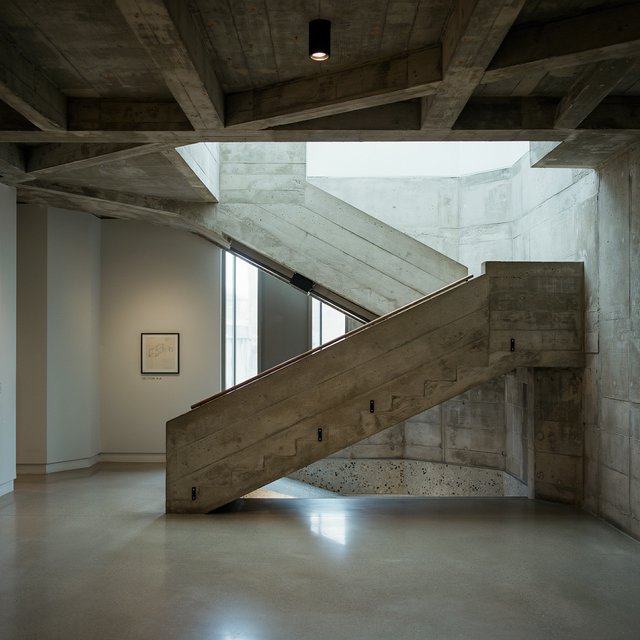At the heart of a restrained Brutalist museum, a folded concrete stair rises beside smoked-oak handrails and terrazzo landings; skylight washes the treads, seen from the lower landing, with aligned flights and believable supports. — Ideogram v4