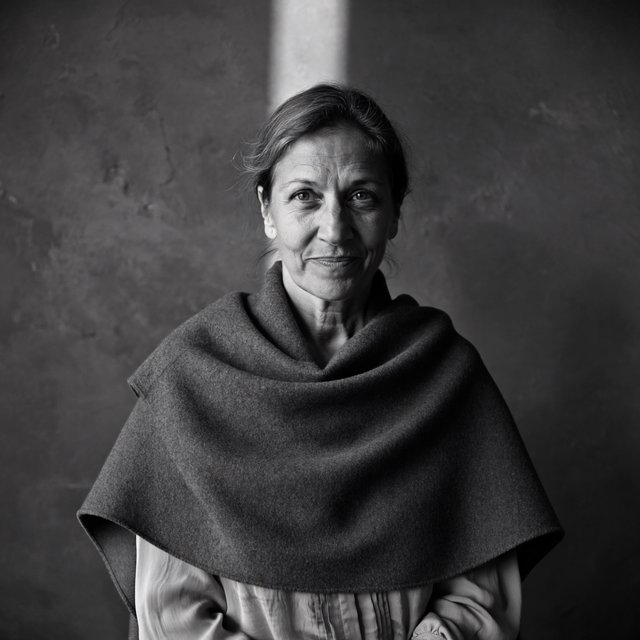 Under a narrow window beam, an adult woman in her late 50s wears a sculpted wool cape over a cotton blouse; her wry expression rests against a charcoal plaster backdrop in a black-and-white portrait, shaped by soft angled light. — ImagineArt 2