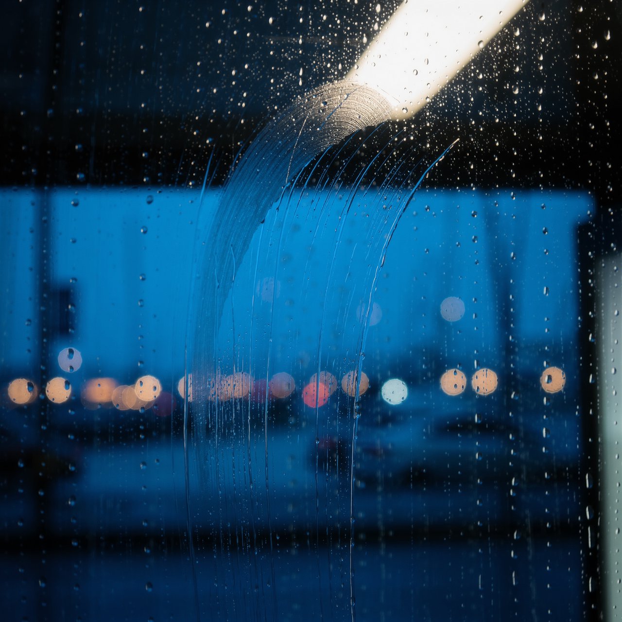 Raindrops crawl down a bus shelter pane, warping blurred harbor lights beyond. A handheld evening street snapshot in cool sodium glow, shot at 50mm with shallow depth of field, catches tiny water trails and a faint wipe mark. — Kling v3 Omni