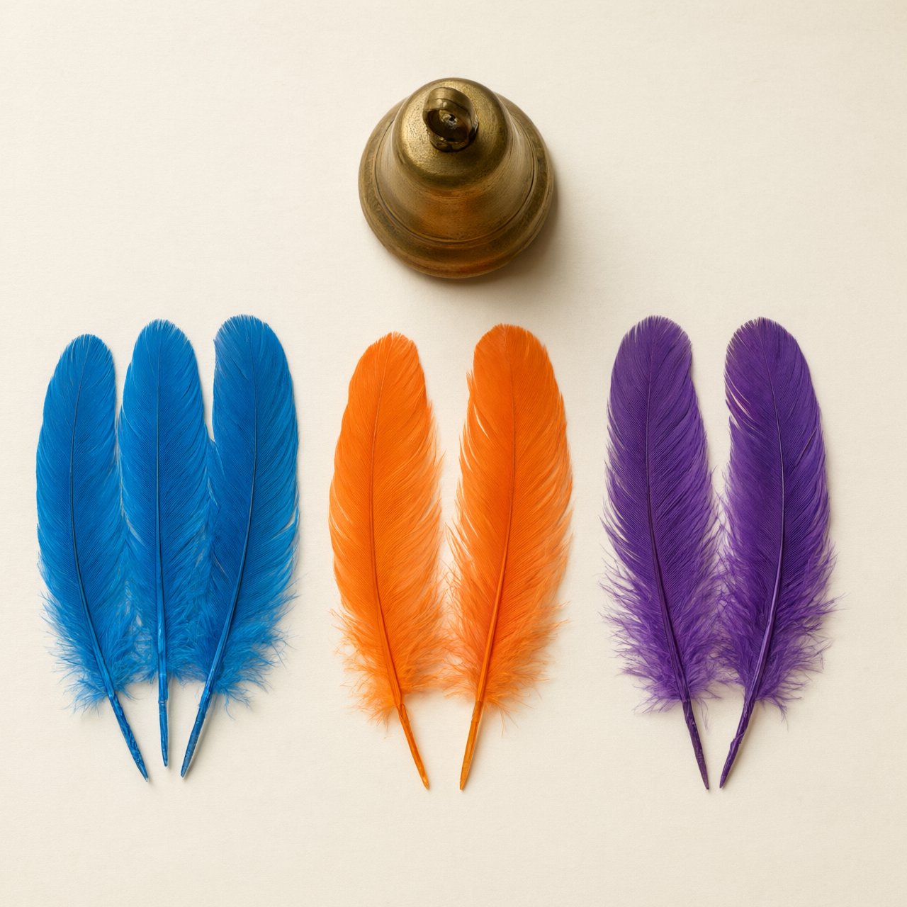 Exactly seven feathers lie on a cream tabletop: three blue on the left, two orange in the center, and two purple on the right. A brass bell sits above the orange pair, nothing else. — Qwen Image 3.0 Pro
