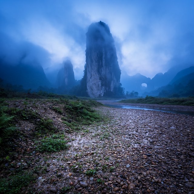 A limestone karst valley at blue hour after a light shower, towering pale cliffs rising above a clear river, low clouds snagging on pinnacles, slick ferns and rounded pebbles leading toward a distant stone arch. — Krea 2 Large