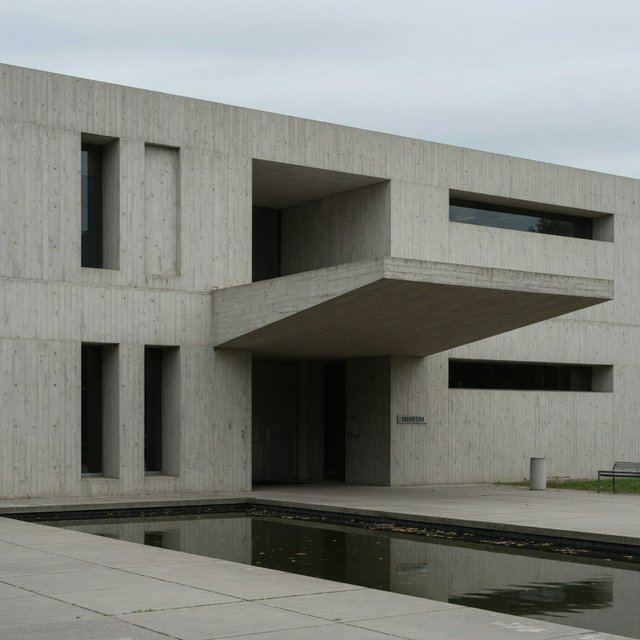 Exterior of a compact brutalist art museum, pale board-formed concrete, deep rectangular window recesses, a broad cantilevered entrance canopy, low reflecting pool, overcast afternoon light, viewed from a quiet plaza at eye level, crisp orthogonal architectural photography. — Grok Imagine Image Quality
