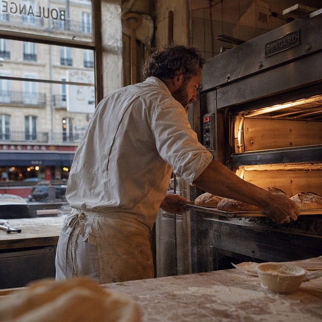 Candid documentary photograph of an adult baker sliding a tray of bread into a neighborhood bakery oven, warm oven glow mixing with gray dawn window light, 35mm lens with moderate depth of field, flour dust suspended above the workbench. — GPT Image 2