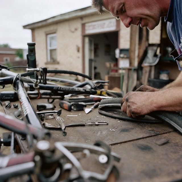 Candid documentary photograph of an adult bicycle mechanic repairing a flat tire outside a neighborhood workshop, soft overcast daylight, 35mm lens with moderate depth of field, greasy fingerprints, dust, and tiny metal shavings visible on the workbench. — ImagineArt 2