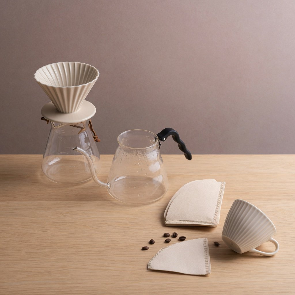 From directly overhead, an unbranded pour-over coffee set with ribbed porcelain and clear borosilicate components arranged in sequence on pale oak; warm taupe background, broad diffused softbox light and gentle side fill shape this commercial product image, no lettering. — Grok Imagine Image Quality