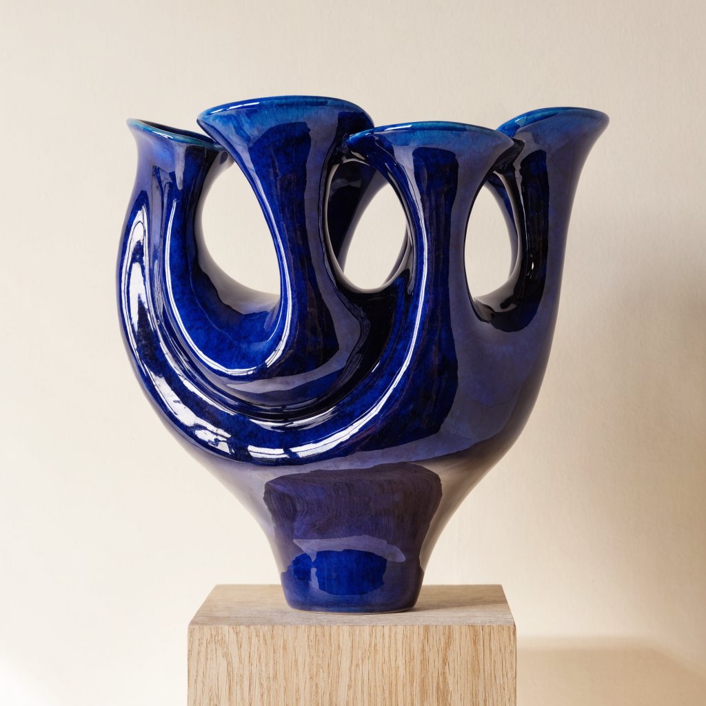 Commercial product photograph of an unbranded sculptural ceramic vase, formed from glossy cobalt porcelain, standing on a pale oak pedestal against a warm ivory background, illuminated by soft directional daylight and subtle fill. — Recraft v4