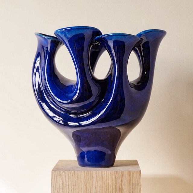 Commercial product photograph of an unbranded sculptural ceramic vase, formed from glossy cobalt porcelain, standing on a pale oak pedestal against a warm ivory background, illuminated by soft directional daylight and subtle fill. — Recraft v4