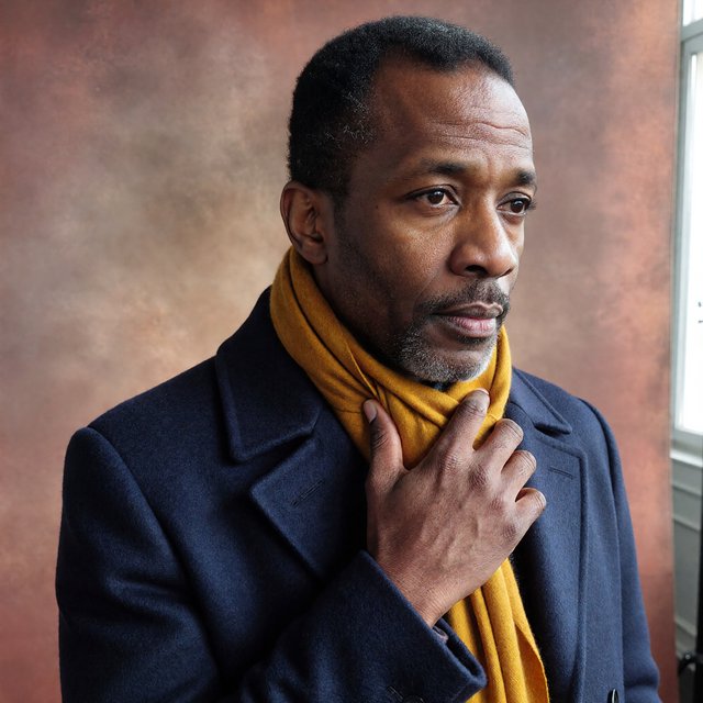 Editorial portrait of an adult Black man in his 30s wearing a deep navy wool overcoat and mustard scarf, fingers adjusting the scarf, thoughtful expression, muted terracotta studio backdrop, crisp cool window light. — ImagineArt 1.5 Pro