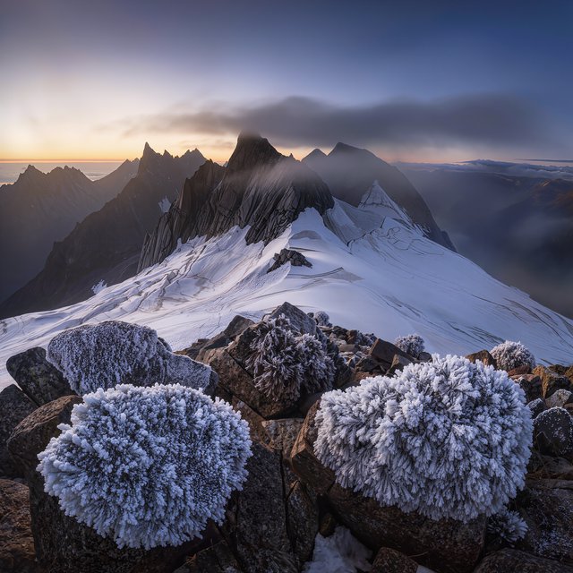 Wind combs a high granite ridge above a glacial cirque at sunrise, beneath clearing snow squalls; granular rime crusts foreground boulders while successive peaks dissolve into blue cloud shadow. — Kling v3