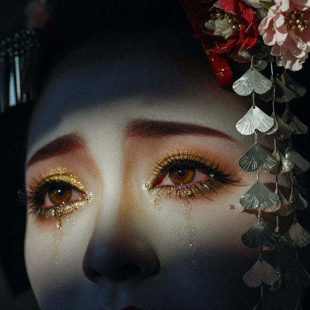 Extreme close-up macro photography of a Geisha's face. The focus is on her eyes, revealing intense emotion and hyper-realistic detail. Her skin is covered in traditional white 'Oshiroi' makeup, showing subtle, realistic cracking and skin texture. Dramatic Chiaroscuro lighting with deep shadows and a single soft light source highlighting the shimmer in her golden eyeshadow and the wetness of her eyes. Ornate kanzashi (hair ornaments) visible in the blurred periphery with intricate silk flowers and dangling silver petals. Cinematic composition, shot on 100mm macro lens, incredibly detailed eyelashes and iris patterns. — Model 6