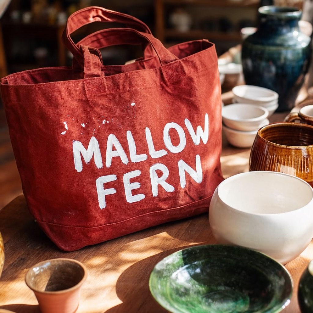 Sunlight pools over a pottery market table, where a rust-red canvas tote rests beside glazed bowls; "MALLOW FERN" is hand-painted in chunky white letters on its front, with no other legible writing. — Krea 2 Large