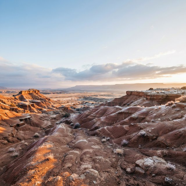 A red-sandstone badlands plateau beneath clearing winter clouds at sunrise, overnight frost silvering the gullies; cracked clay and wind-smoothed rock lead the eye across layered mesas fading into pale distance. — Ideogram v4.5