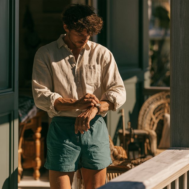 At a sunlit island veranda, an adult man in teal swim shorts and a sand-colored linen shirt is caught fastening his watch, crisp noon light and relaxed tailoring lending fashion-magazine polish. — Midjourney V8.2