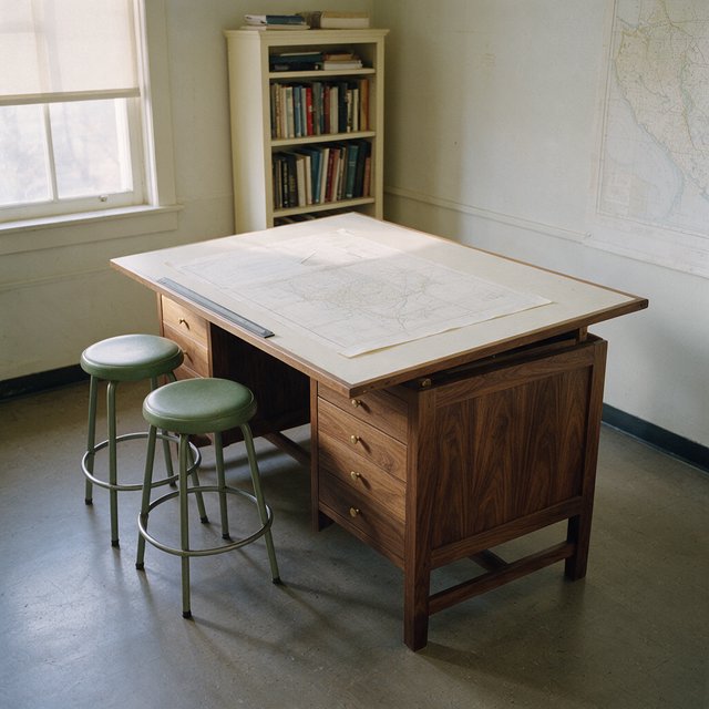 A quiet mapmaking room contains exactly one walnut drafting desk at the center, two moss-green stools side by side along its west edge, and one cream bookcase against the north wall; pale morning light, nothing else. — Seedream 5.0 Pro