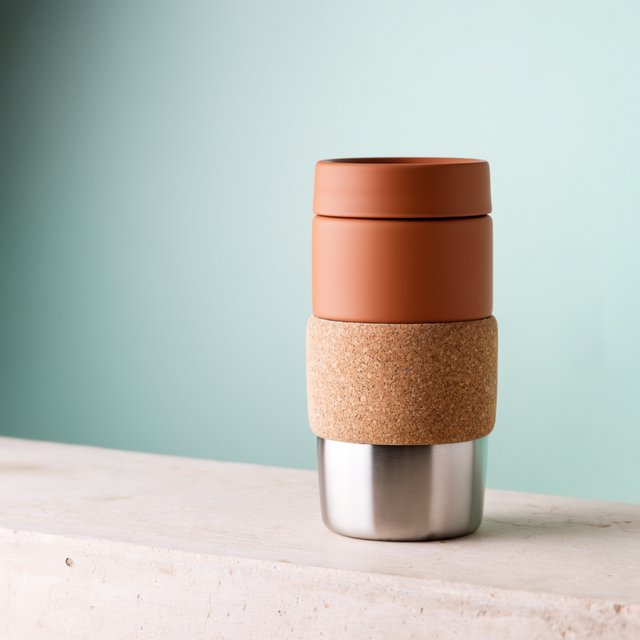 Commercial product photograph of an unbranded insulated travel mug made from matte terracotta powder-coated steel with a cork grip, standing on a pale limestone slab against a soft teal background, lit by broad diffused window light and a gentle edge light. — Krea 2 Large