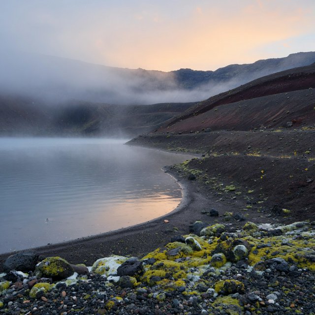 At first light, a rain-cleared volcanic caldera lake lies beneath thinning fog; pumice grains and damp lichen sharpen the near shore, while dark cinder slopes step back toward a distant rim. — Grok Imagine Image Quality