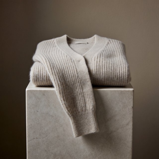 A folded unbranded merino cardigan rests on a honed limestone plinth, its chunky ribbed wool surface softly catching broad overhead diffusion and warm side bounce against a muted clay backdrop, with no tags, lettering, or graphics. — Krea 2 Medium
