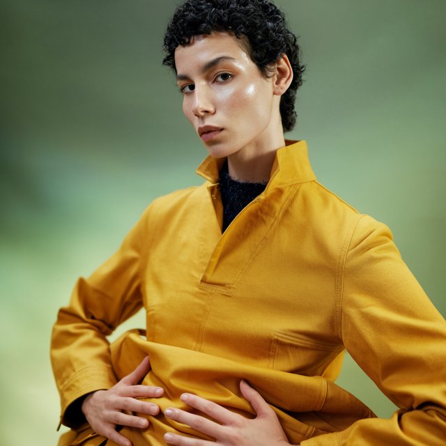 Fashion portrait of an adult nonbinary person in their late twenties wearing a structured saffron-yellow canvas jacket over a fine-gauge navy merino top, hands resting on the jacket hem. Their bright, self-assured expression contrasts with a softly lit sage-green backdrop. — Krea 2 Large