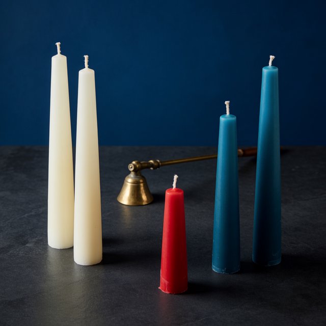 Exactly five candles stand on a dark slate tabletop: two ivory candles on the left, one red candle in the center, and two teal candles on the right. A brass snuffer sits above the red candle, nothing else. — Krea 2 Medium